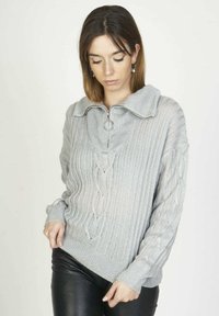 Mujer con cabello castaño lacio que lleva pendientes de estrellas colgantes, un suéter de punto acanalado gris con cremallera y cuello, y pantalones de cuero negros.