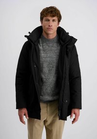Schwarze Daunenjacke mit hohem Kragen, Reißverschluss und Druckknöpfen. Trägt sie über einem grauen Pullover. Mann steht vor einem neutralen Hintergrund.