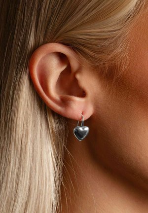 Silberner herzförmiger Ohrring an einem durchstochenen Ohr, mit hohem Glanz und glatter Textur. Das Haar ist glatt gestylt und blond.