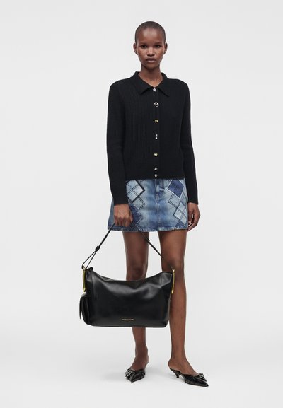 Femme portant un cardigan noir boutonné et une jupe en denim bleue à motifs, tenant un grand sac à main noir Marc Jacobs par les poignées.