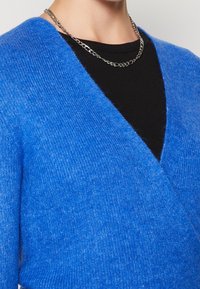 Sininen pörröinen kietaisuneule mustan ribatulla paidan päällä, kaulassa hopeaketju, paljastaen ylärinnan ja kaulan alueen.