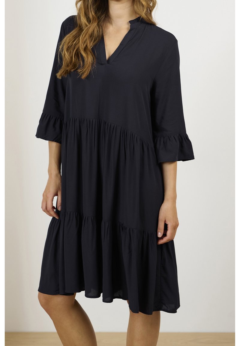 Femme portant une robe noire ample mi-longue à manches trois-quarts avec des détails à volants superposés, debout contre un mur uni.