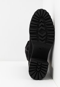 Semelle de botte noire avec des crampons profonds et à motifs, placée verticalement contre un coin de mur et de sol blancs.
