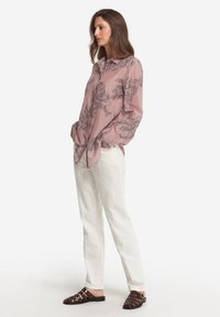 Långärmad, ljusrosa blus med svart blommönster; lös passform. Kombinerad med vita rakbenade byxor och vävda, mörkbruna sandaler.