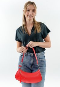 Borsa a tracolla in pelle rossa con forma curva, struttura in metallo argentato e un charm decorativo a forma di croce. Indossata con un outfit casual e jeans di denim.