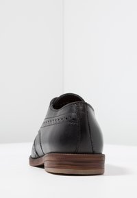 Černá kožená bota s hladkým povrchem, zdobená perforacemi a hnědou gumovou podrážkou. Zadní pohled zdůrazňuje zakřivený podpatek.