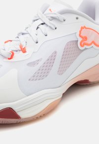 Scarpa sportiva con una tomaia in rete grigia chiara, accenti bianchi, dettagli arancioni e una suola semi-trasparente con una sezione rossa.