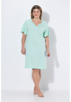 Žena se světle hnědými vlasy stojí bosá, oblečená v volných šatech délky ke kolenům v mátově zelené barvě s krátkými volánovými rukávy a výstřihem se zářezem.