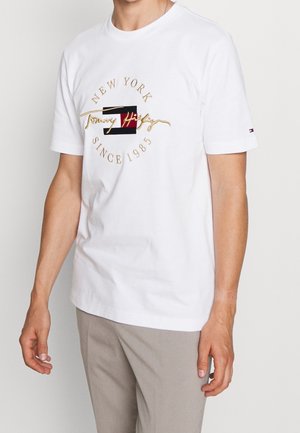 White cotton t-shirt featuring a gold embroidered logo with "New York" and "Tommy Hilfiger" text, flanked by a red and black rectangular design.