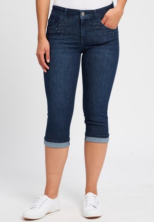 Dunkelblaue Denim-Capri-Jeans mit umgeschlagenen Säumen, vorderen Taschen mit Nietenakzenten und einem klassischen Knopfverschluss. Kombiniert mit weißen Sneakers.