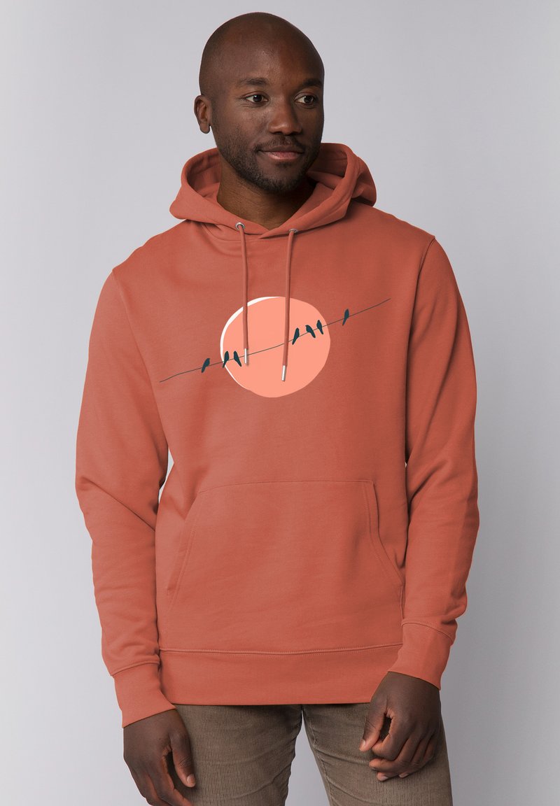 Roestkleurige hoodie met een kangoeroezak, voorzien van een grafisch ontwerp van gesilhouetteerde vogels op een draad tegen een cirkelvormige zon als achtergrond.