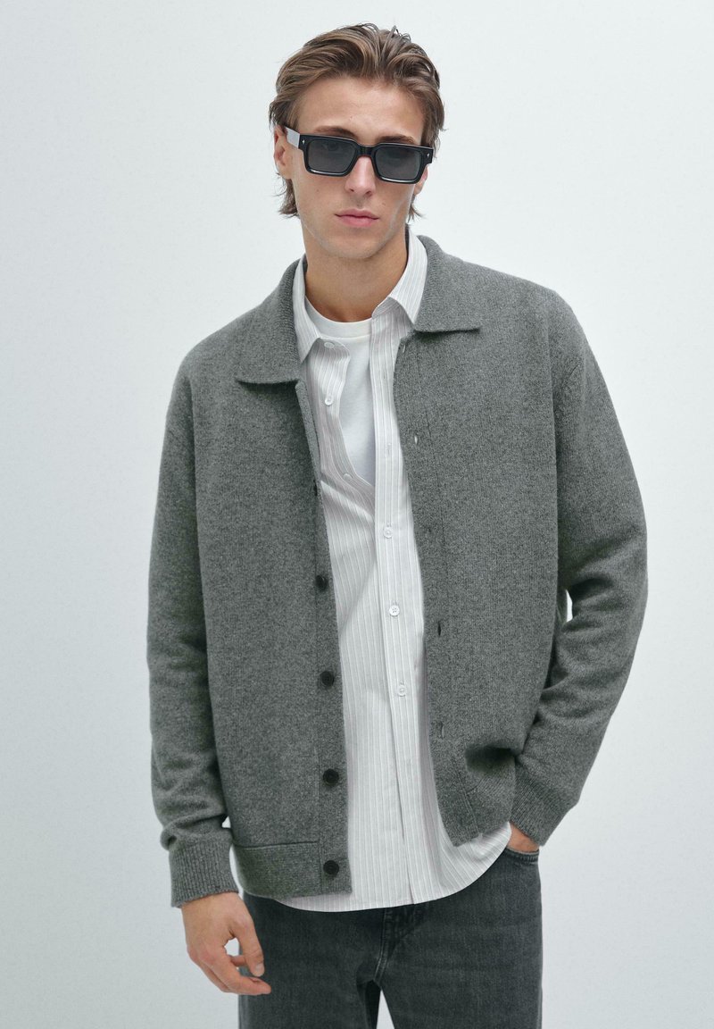 Giovane uomo con occhiali da sole scuri, cardigan grigio con bottoni sopra una maglietta bianca a righe e jeans, con una mano in tasca su uno sfondo semplice.