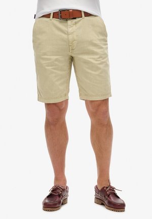 Bermudas beiges con un tejido texturizado, que presentan un cierre de botón y trabillas para el cinturón, combinadas con zapatos de cuero marrón.