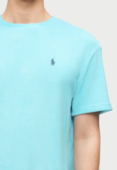 Άτομο που φοράει ένα ανοιχτό μπλε t-shirt με κοντά μανίκια και μικρό λογότυπο σκοτς παίκτη πόλο σε σκούρο μπλε χρώμα στο αριστερό στήθος.