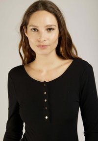 Haut à manches longues noir à côtes, avec un encolure dégagée et cinq boutons décoratifs au centre. Texture douce avec un léger stretch.
