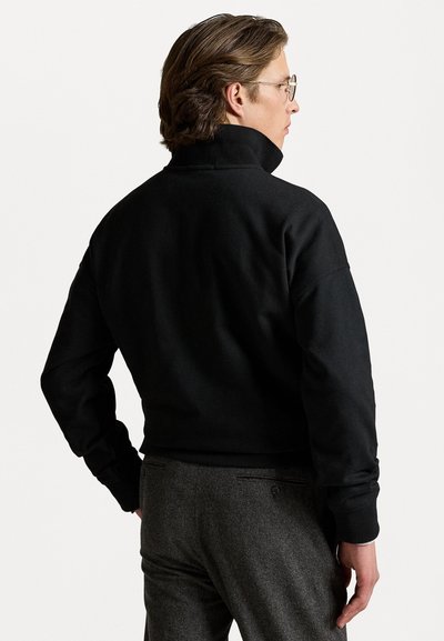 Μαύρο φούτερ με ψηλό γιακά και ριμπ μανσέτες, συνδυασμένο με γκρι υφηρό παντελόνι. Η πίσω όψη δείχνει άνετη εφαρμογή και καθαρές γραμμές.