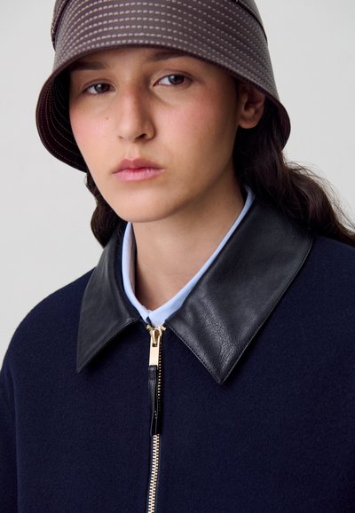 Chaqueta navy con cremallera y cuello de cuero negro, combinada con un cuello de color azul claro debajo y un sombrero de cubo marrón con patrón.