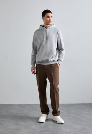 Šedá mikina s kapucí a přední kapsou, na šňůrku, s modrým logem. Nosená s hnědými kalhotami a bílými teniskami na hladkém šedém povrchu.