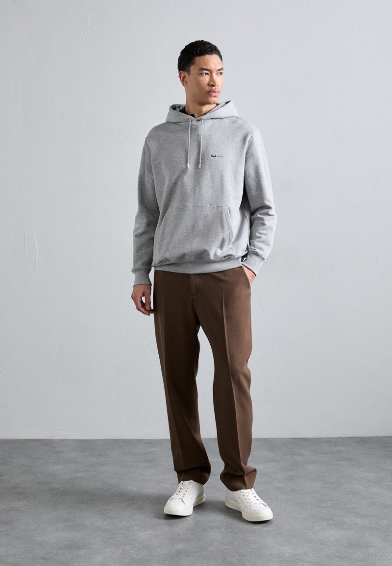 Siva dukserica s kapuljačom i prednjim džepom, s plavim logotipom. Nosena je uz smešane hlače i bele patike na glatkoj sivoj podlozi.