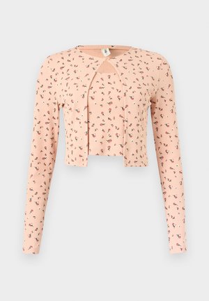 Jonge persoon draagt een perzikkleurige, bloemrijke, korte cardigan en top, losse zwarte jeans, zwart-witte sneakers, en houdt een donkere clutch vast.