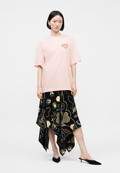 Γυναίκα που φοράει φαρδιά ροζ μπλούζα με λεπτομέρεια καρδιάς και μαύρη ασύμμετρη φλοράλ φούστα, στέκεται με το ένα πόδι λίγο πιο μπροστά.