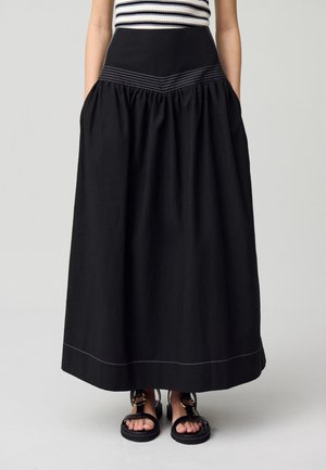 Larga falda negra con un diseño fruncido, que cuenta con una cinta en la cintura ajustada y costuras blancas en contraste. Se lleva con sandalias negras.