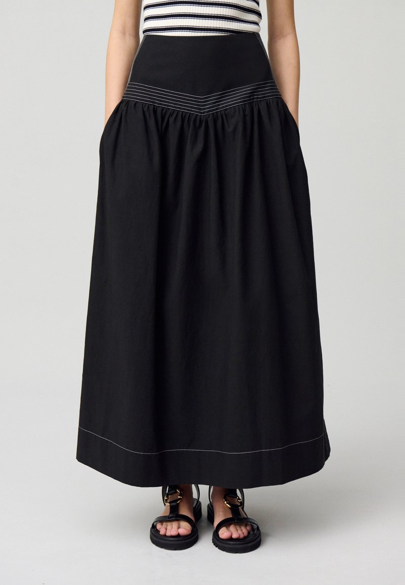 Jupe longue noire avec un design froncé, dotée d'une taille ajustée et d'une couture blanche contrastante. Associée à des sandales noires.