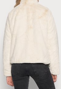 Chaqueta de piel sintética color crema con cuello alto, corte relajado y texturización suave. Presenta un color sólido y sutiles variaciones en el tono.