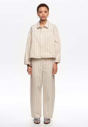 Joven con chaqueta de raya vertical color crema y pantalones anchos a juego, con sandalias grises abiertas, de pie contra un fondo blanco.