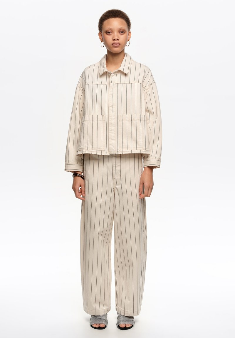 Mladá osoba oblečená v krémovom blejzri s vertikálnymi tenkými pásikmi a zodpovedajúcich širokých nohaviciach, s otvorenými sivými sandálmi, stojaca pred bielym pozadím.