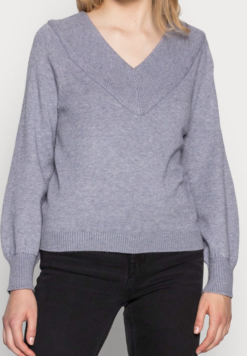Femme portant un pull en maille léger violet clair à col en V avec poignets et ourlet côtelés, associé à un pantalon noir.