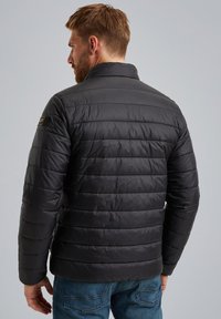 Chaqueta de plumas negra con diseño de acolchado horizontal, cuello alto y detalle del logo en el hombro izquierdo, combinada con jeans azules.