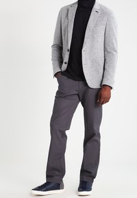 Hombre con blazer gris, suéter de cuello alto negro, pantalones gris oscuro y zapatillas de deporte azul marino, de pie con las manos en los bolsillos.