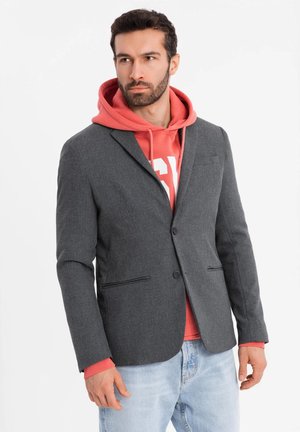 Grauer Blazer aus strukturiertem Stoff, mit einem Knopfverschluss, zwei frontalen Taschen, über einem roten Hoodie getragen. Lässiger Stil.
