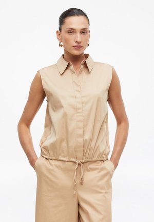 Vrouw met haar naar achteren gebonden, draagt een mouwloos beige overhemd met kraag en bijpassende beige broek met trekkoord, handen in de zakken.