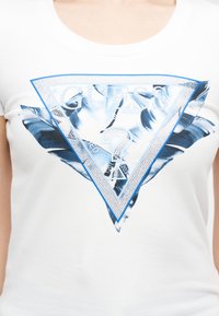 Κοντινή λήψη λευκού T-shirt με σχέδιο μπλε φύλλου και ανακλαστικό ασημένιο τρίγωνο με το λογότυπο "Guess" και ένα ερωτηματικό.