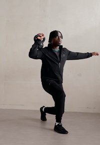 Schwarze Sportbekleidung, einschließlich einer Reißverschlussjacke und Jogginghosen, mit einer über dem Kopf gehaltenen Kettlebell von einer Person in einer dynamischen Haltung vor einem neutralen Hintergrund.