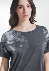 Ung kvinna med vågigt brunt hår som bär en urtvättad svart T-shirt med abstrakta vita och grå grafiska mönster över axlarna.