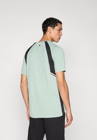 Sportshirt in muntgroen met zwarte accenten, korte mouwen en een ronde halslijn. Heeft een contrasterende streep aan de achterkant.