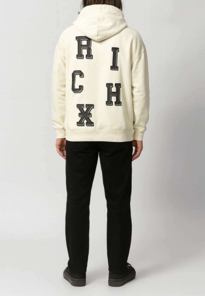 Felpa cremosa con lettere ricamate nere "RICH" e "CHX" in diversi caratteri sulla schiena. Abbinata a pantaloni neri e scarpe grigie.