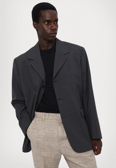 Tumedast halli bleiser, mis on valmistatud tekstuurist kangast, millel on nurkadega krae, üks rinnatasku ja nööbiga kinnitamine, koos heledate mustriga pükstega.