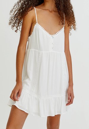 Femme portant une robe blanche sans manches avec de fines bretelles, un col en dentelle, des boutons à l’avant et un ourlet à volants.