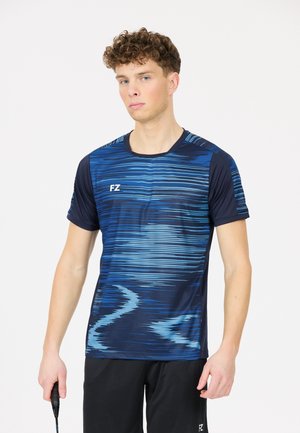 Nuori mies sinisessä kuvioidussa urheilupaidassa ja mustissa shortsissa pitelemässä sulkapallomailaa valkoisella taustalla.