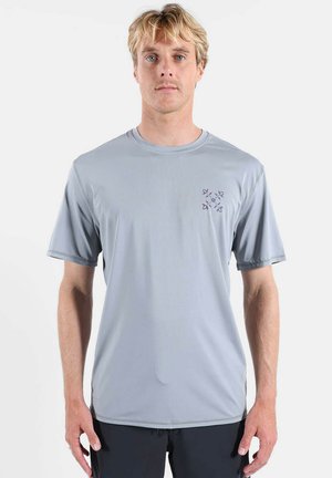 Homme blond debout face à l’avant portant une chemise à manches courtes gris clair avec un petit logo géométrique violet sur la poitrine gauche.