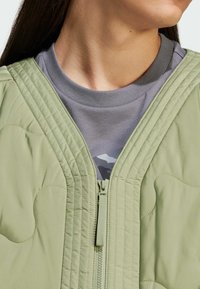 Zelena prešita jakna z zadrgo in rebrastim ovratnikom, nosena čez sivo majico. Ima teksturirano tkanino in subtilen vzorec.