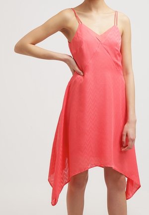 Femme portant une robe corail sans manches avec de fines bretelles doubles et un ourlet asymétrique, debout avec une main sur la hanche.