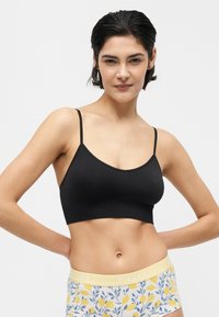 Γυναίκα με κοντά σκούρα μαλλιά, που φοράει μαύρο cropped top και κίτρινα σορτς με λουλουδάτο σχέδιο, με τα χέρια στη μέση, φωτεινό φόντο.