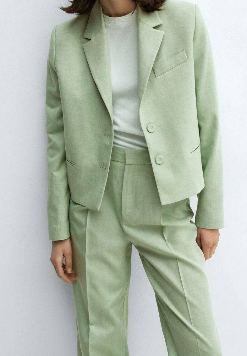 Femme portant un costume vert clair sur mesure avec une veste courte et un pantalon taille haute sur un haut blanc, debout devant un fond clair.