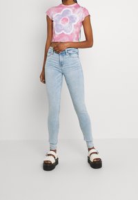 Růžový tie-dye top na krátké rukávy s velkým grafickým motivem fialové květiny, kombinovaný se světle modrými skinny džíny a bílými platformovými sandály s černými podrážkami.