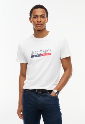 Άνδρας φοράει λευκό t-shirt Tommy Jeans με λογότυπο, σκούρο μπλε τζιν, μαύρη ζώνη και ασημένιο ρολόι χειρός σε απλό λευκό φόντο.